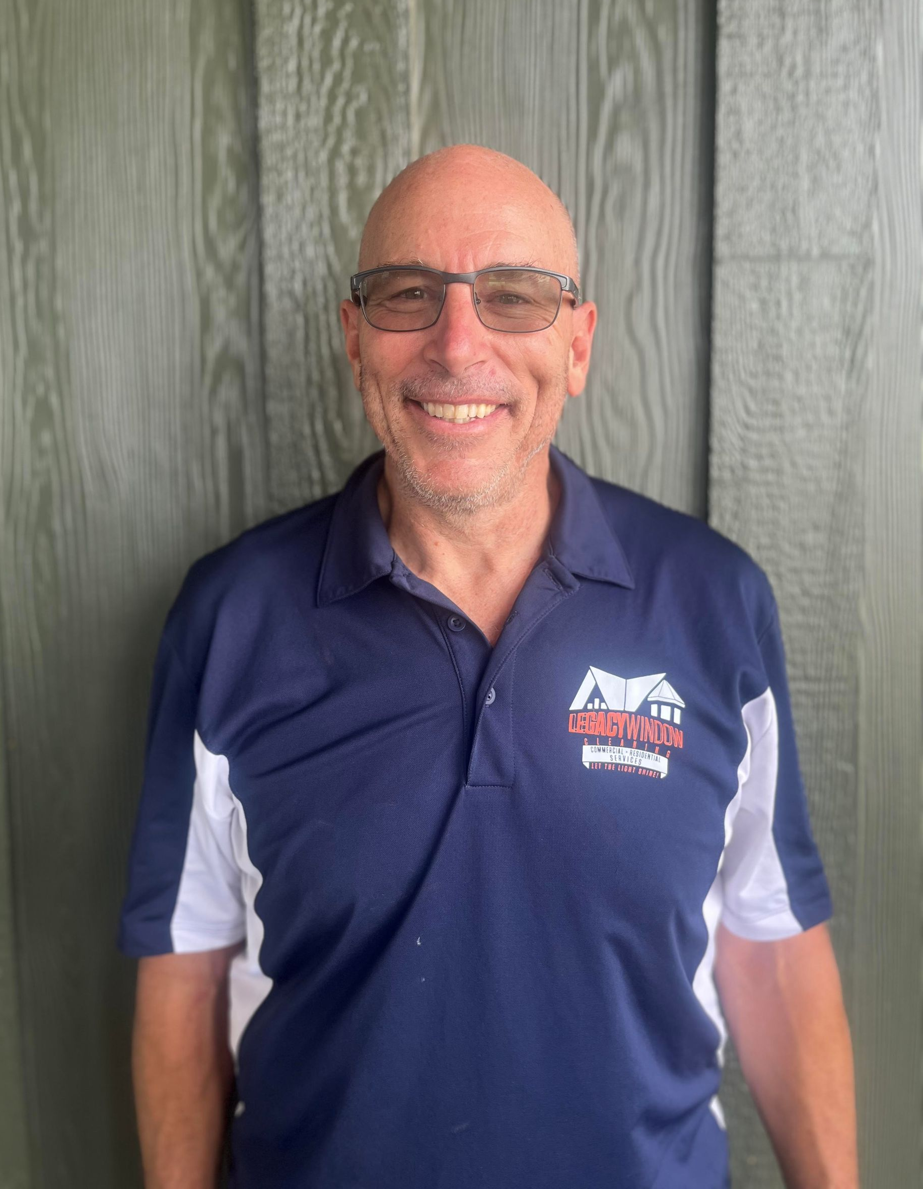 A bald man wearing glasses and a blue shirt is smiling in front of a wooden wall.