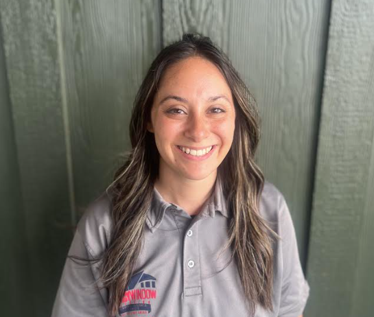 A woman in a grey shirt is smiling in front of a green wall.