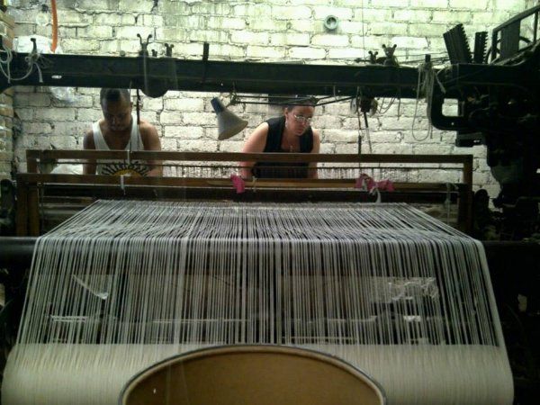 Two women are working on a loom in front of a brick wall