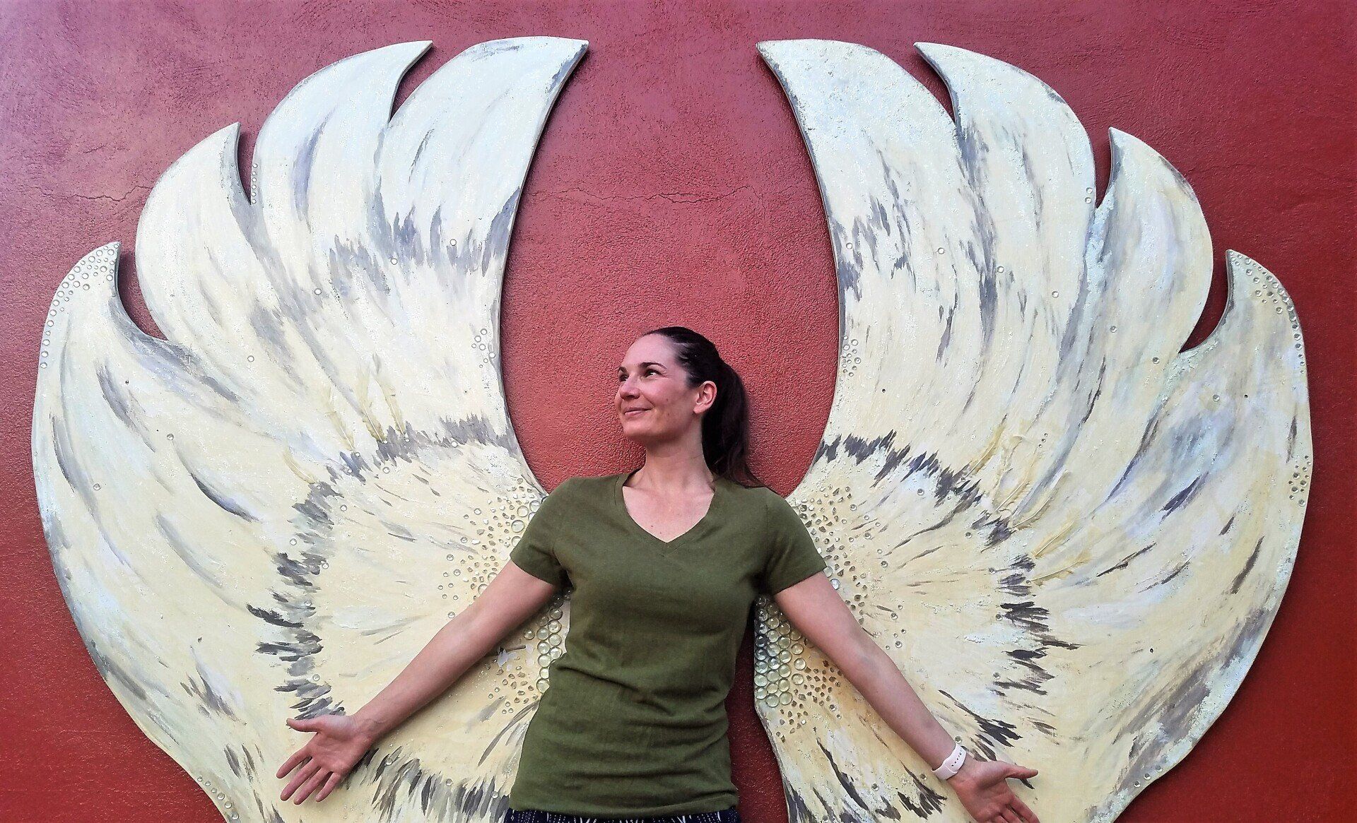 A woman is standing in front of a large angel wing on a red wall.