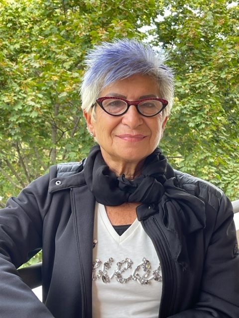 Persona con capelli corti color argento e occhiali rossi, sorridente all'aperto in mezzo alla vegetazione.