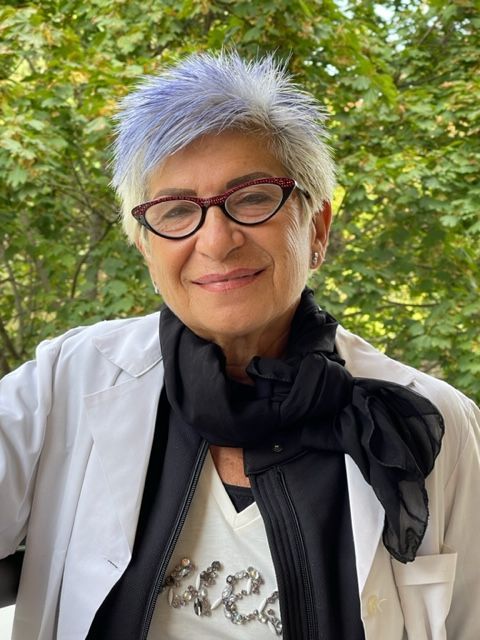 Persona sorridente con capelli corti grigi e occhiali, all'aperto in mezzo al verde.