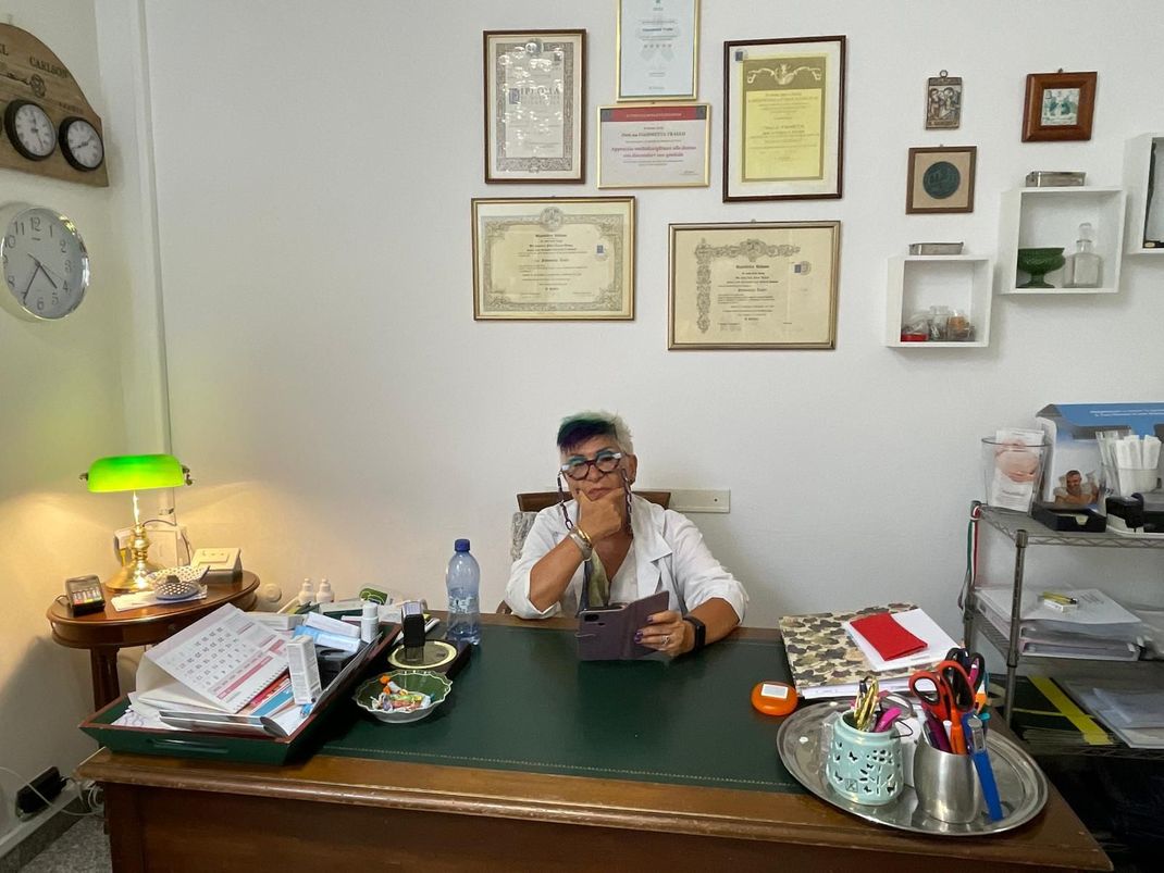 Persona seduta a una scrivania in ufficio, circondata da certificati incorniciati e lampade da scrivania.