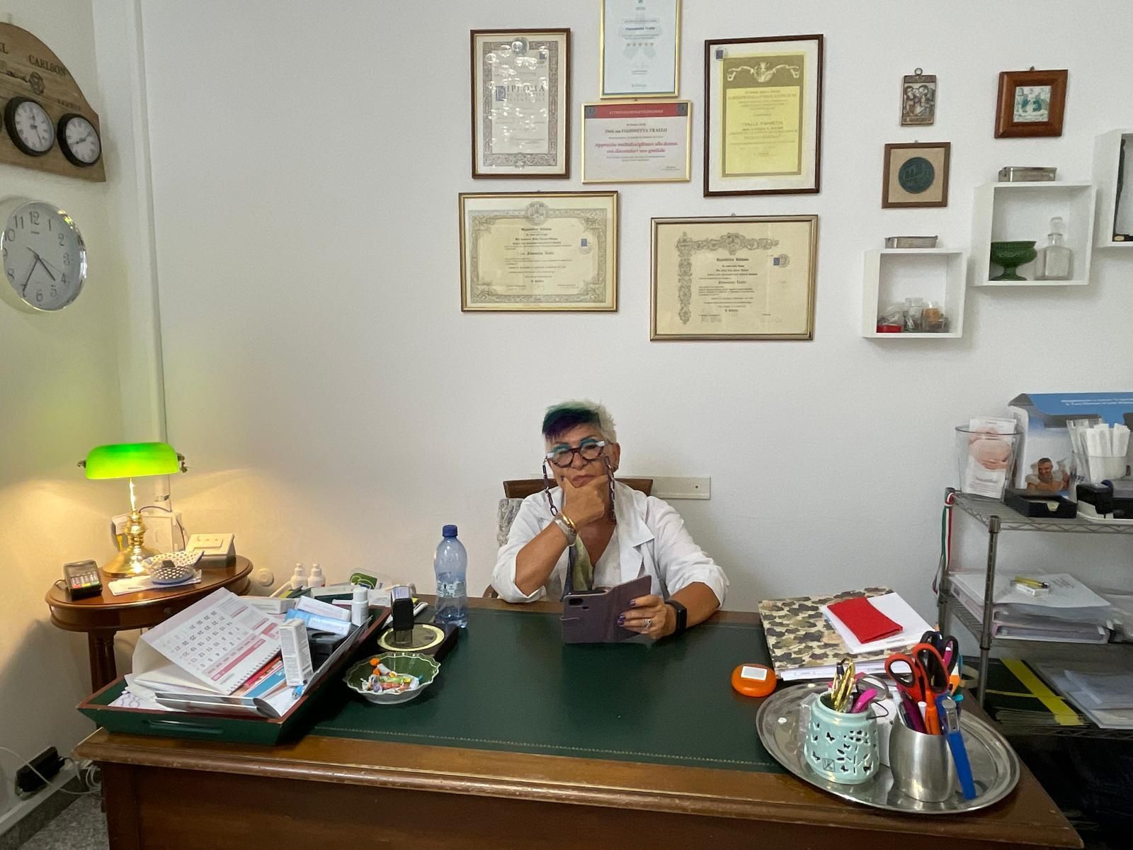 Persona seduta a una scrivania in un ufficio, con certificati incorniciati appesi al muro e carte sparse intorno.