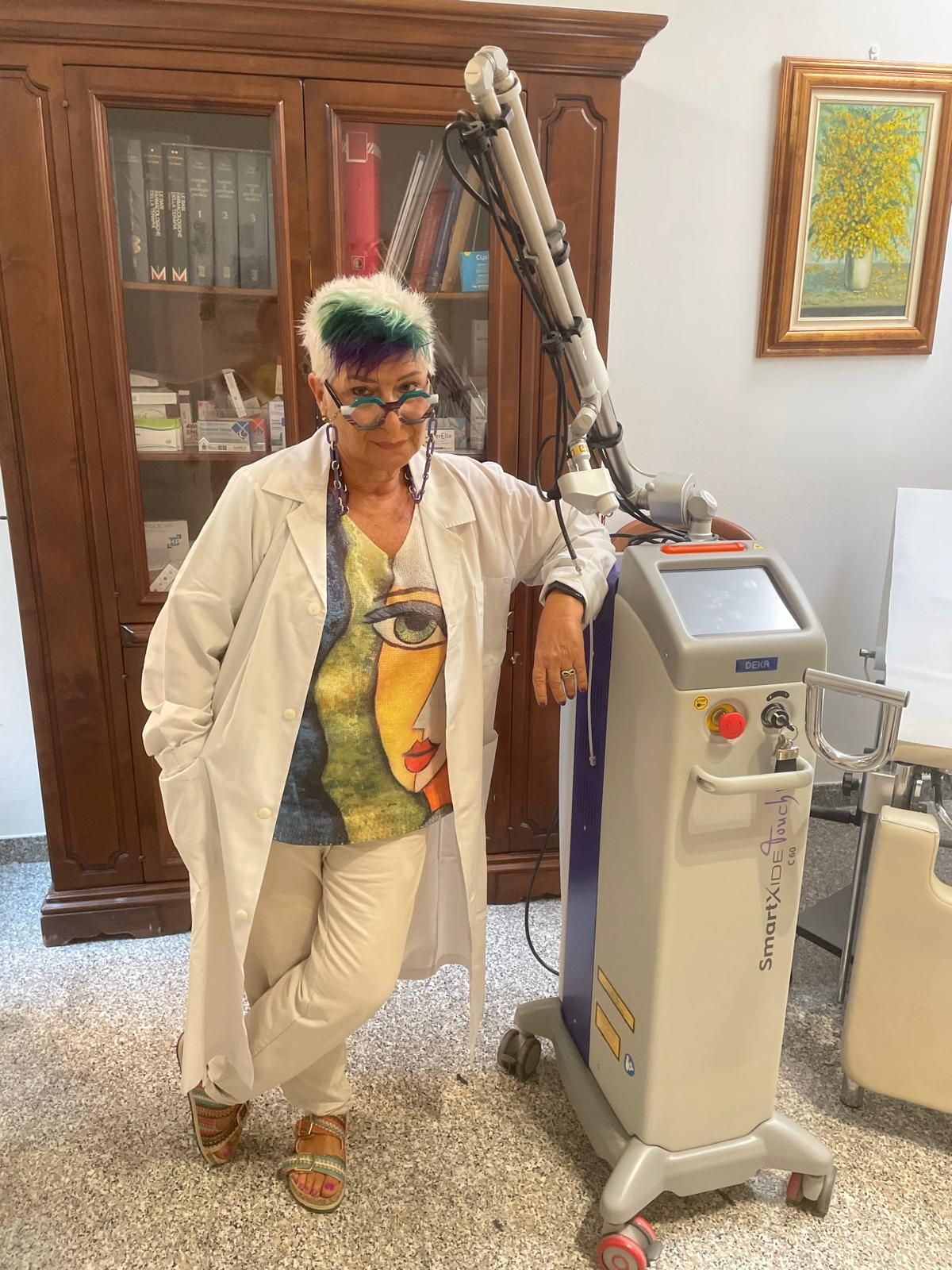 Persona appoggiata a un dispositivo medico in un soggiorno, con indosso abiti bianchi e occhiali colorati.