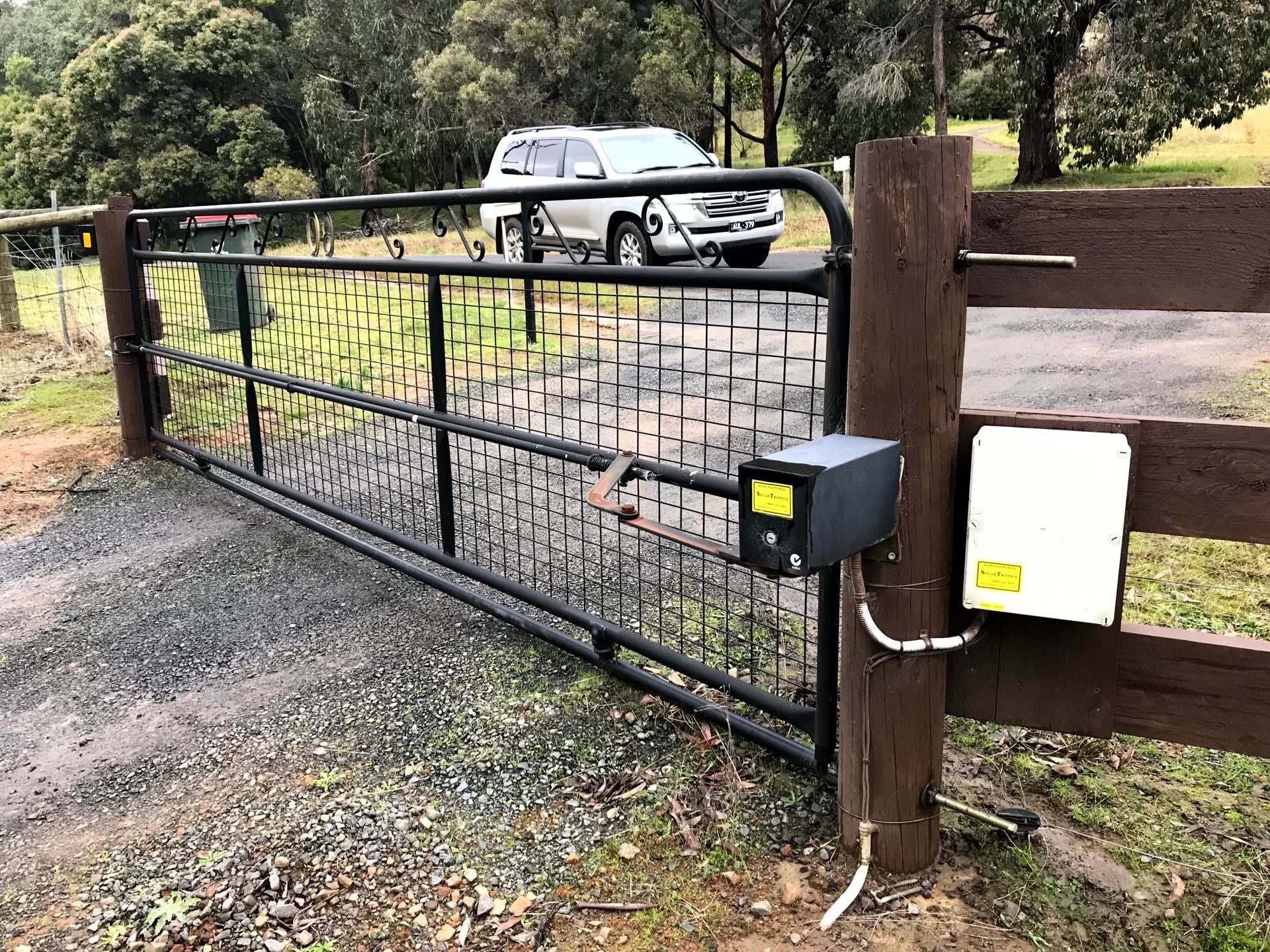Black metal farm gate with keypad beside gravel driveway and parked SUV in the background
