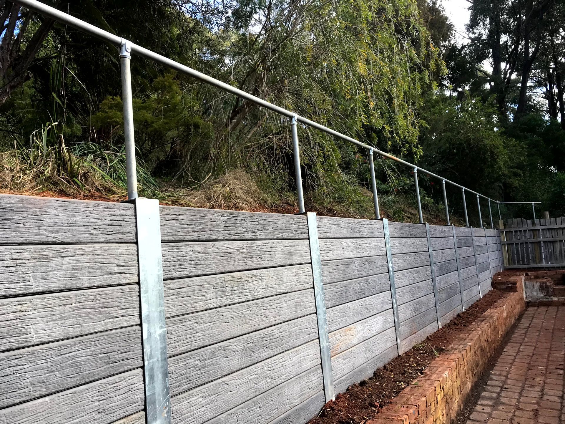 Concrete retaining wall with metal railings beside a brick walkway and trees above