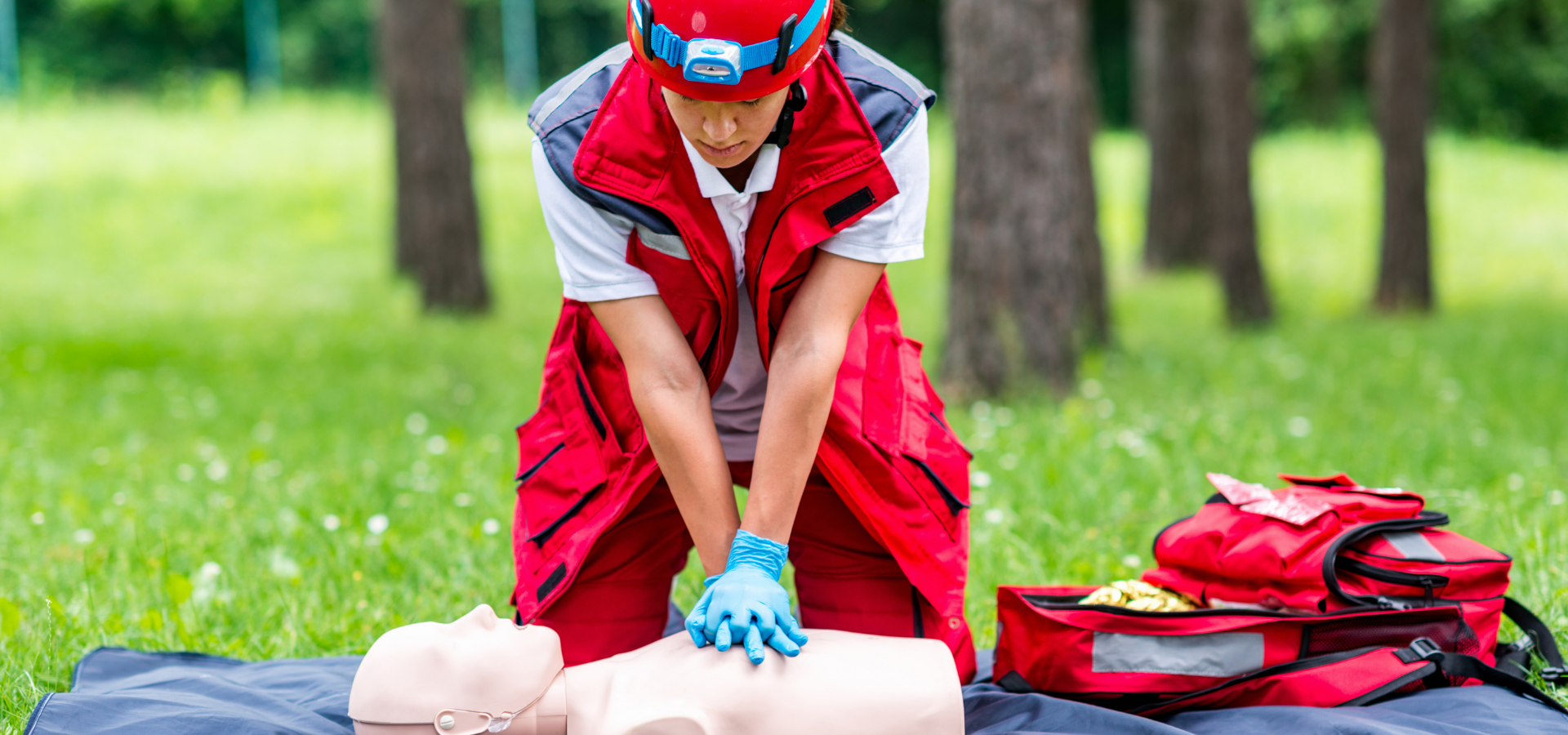 Rescuer in red uniform performing CPR on a manikin outdoors beside an open emergency kit
