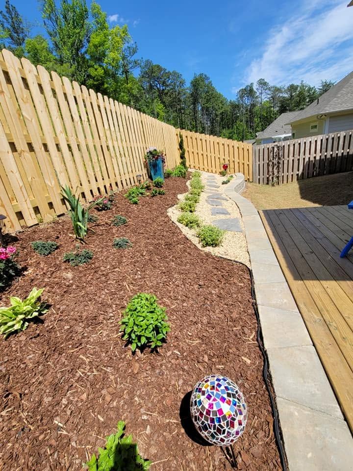 Curved landscaped garden bed beside a wooden deck and tall fence, with small plants and colorful garden orbs.
