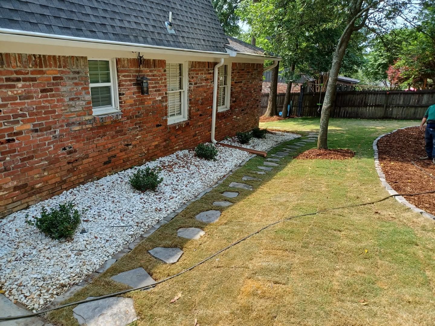 Brick house side yard with white gravel bed, stepping stones, and a tree beside a lawn