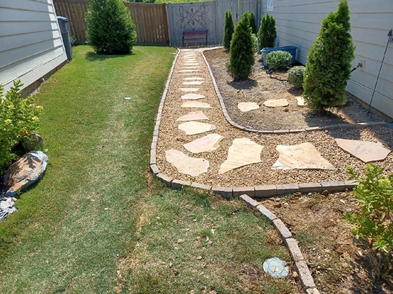 Backyard with a stone path, mulch beds, and small evergreen shrubs along a fence.