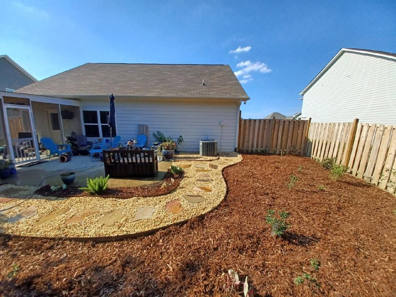 Backyard with fresh mulch, a curved stone border, patio chairs, and a wooden fence under a blue sky