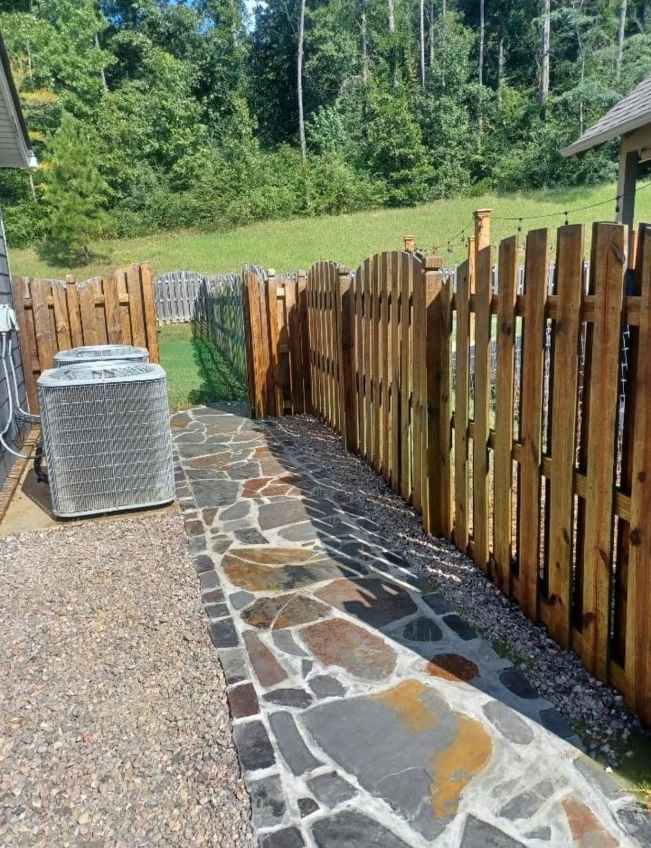 Stone path beside wooden fence in a sunny backyard, with gravel and a wicker-covered table.