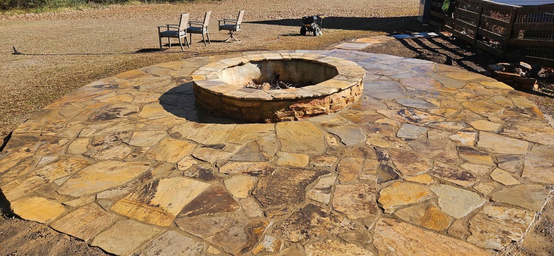 Stone patio with a circular fire pit in a dry outdoor yard, with benches in the background