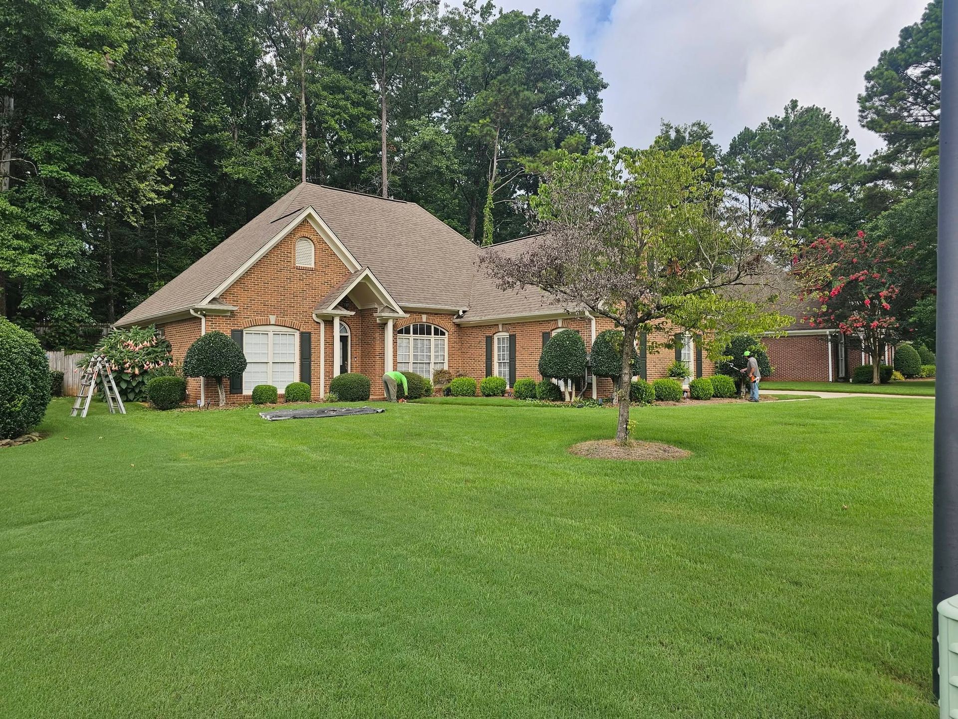 Brick house with a steep roof, surrounded by a large green lawn and trees under a cloudy sky