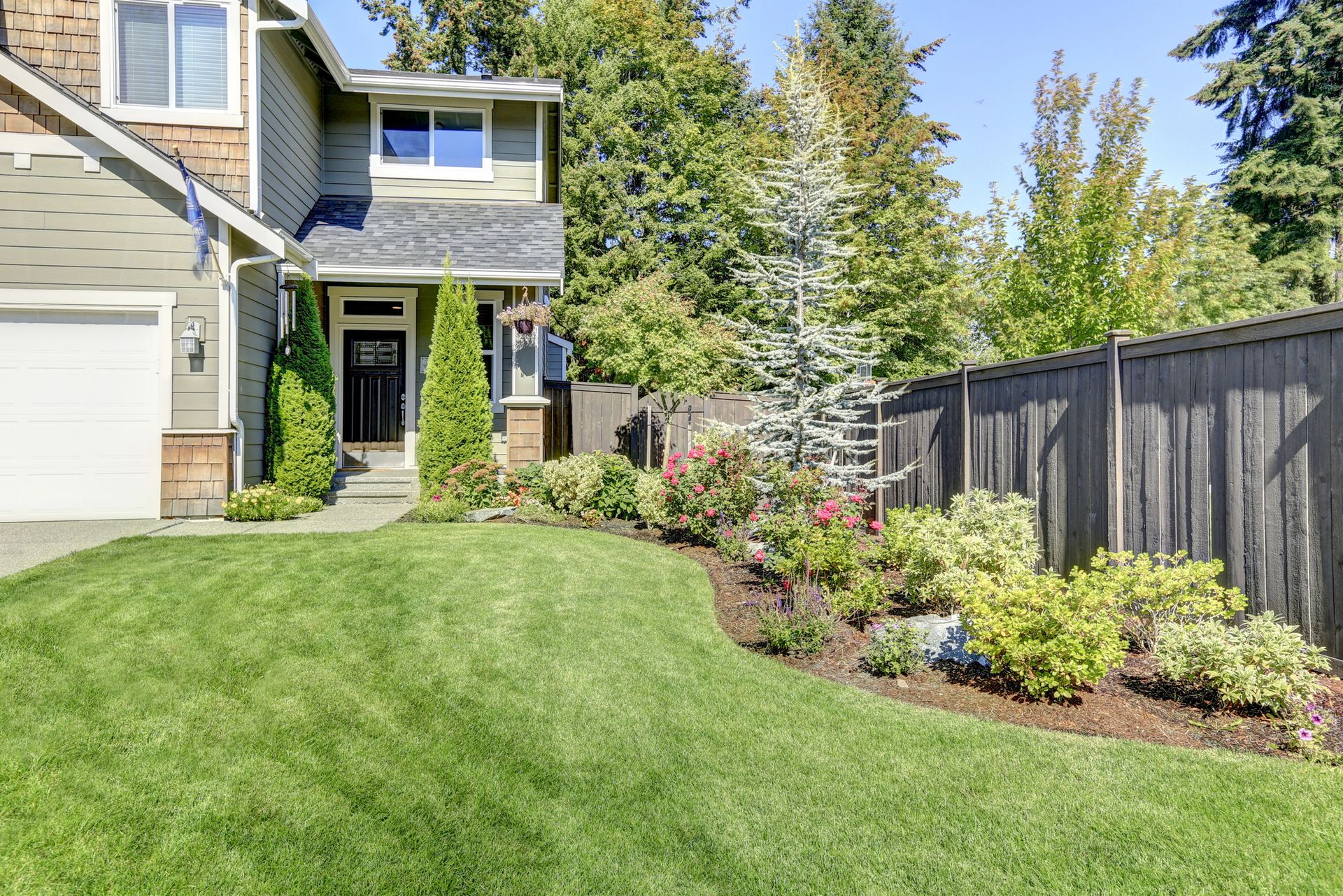 Front yard with green lawn, flower beds, and a house entrance beside a wooden fence.
