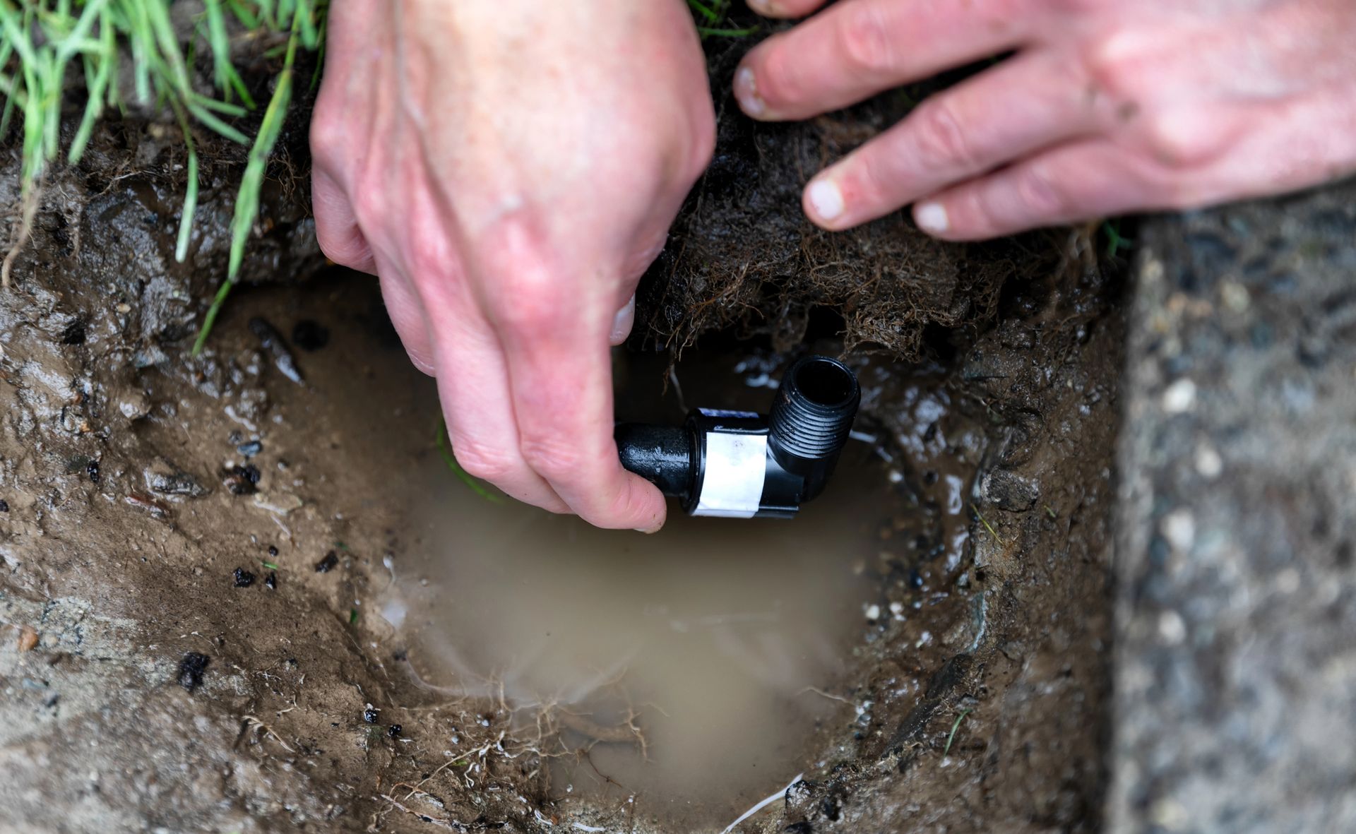 Hands install a black pipe fitting in a muddy trench with water pooled below.