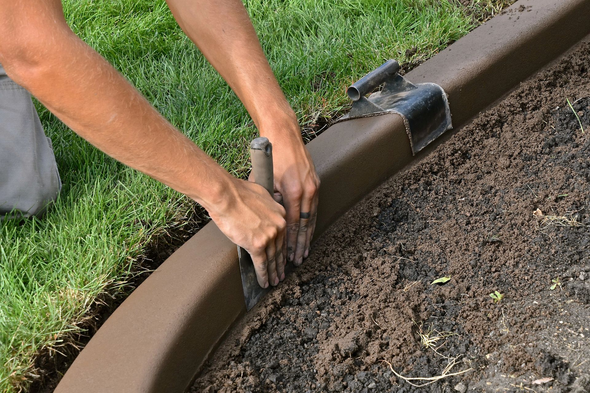 Hands edging a garden bed with a metal edging tool beside soil and grass