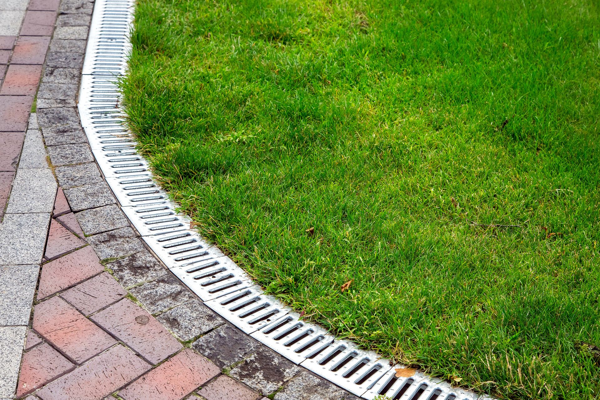 Curved brick path beside a green lawn with a white drainage grate along the edge