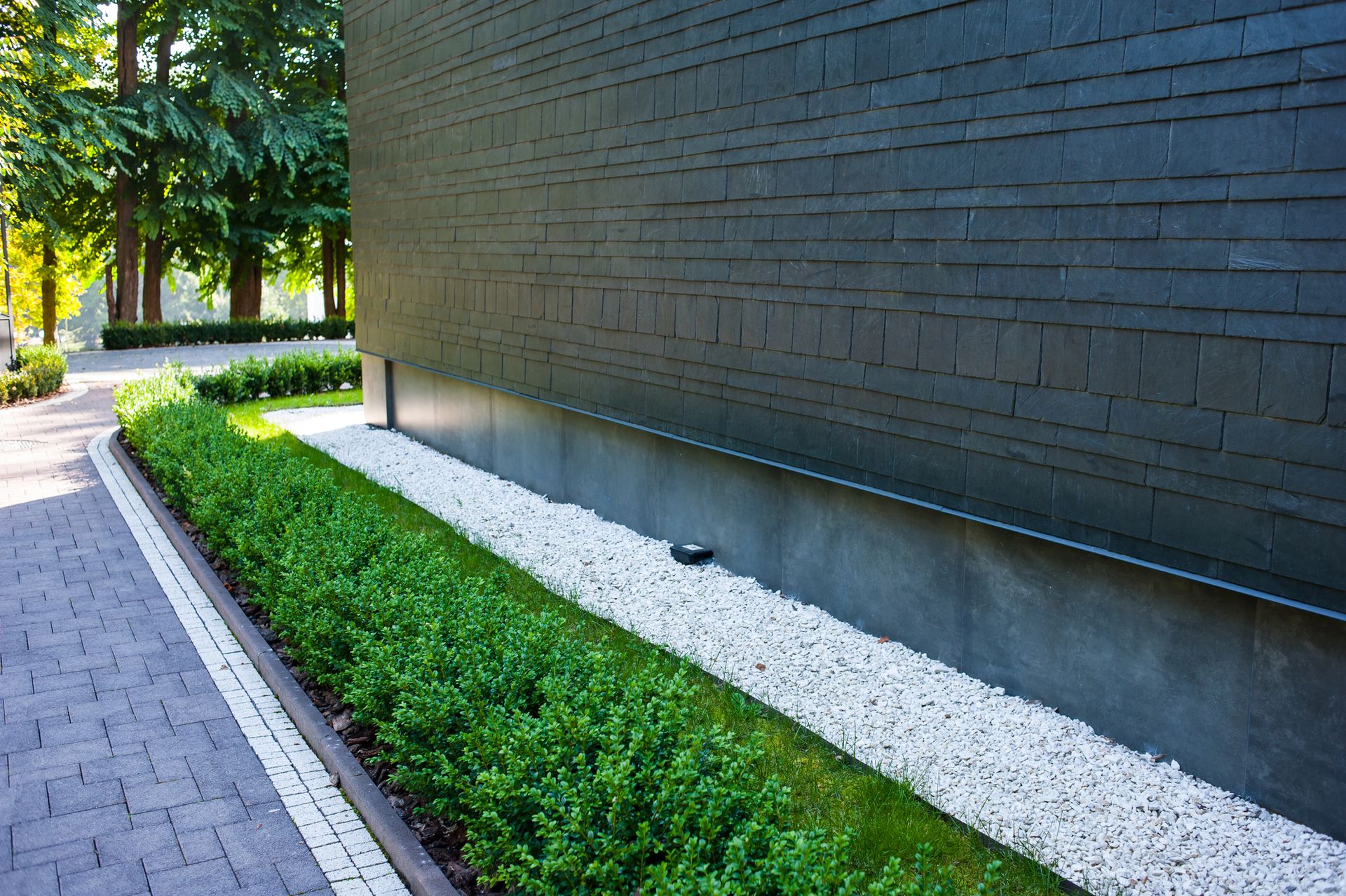 Modern building wall with a narrow white gravel bed, green shrubs, and a paved walkway beside trees