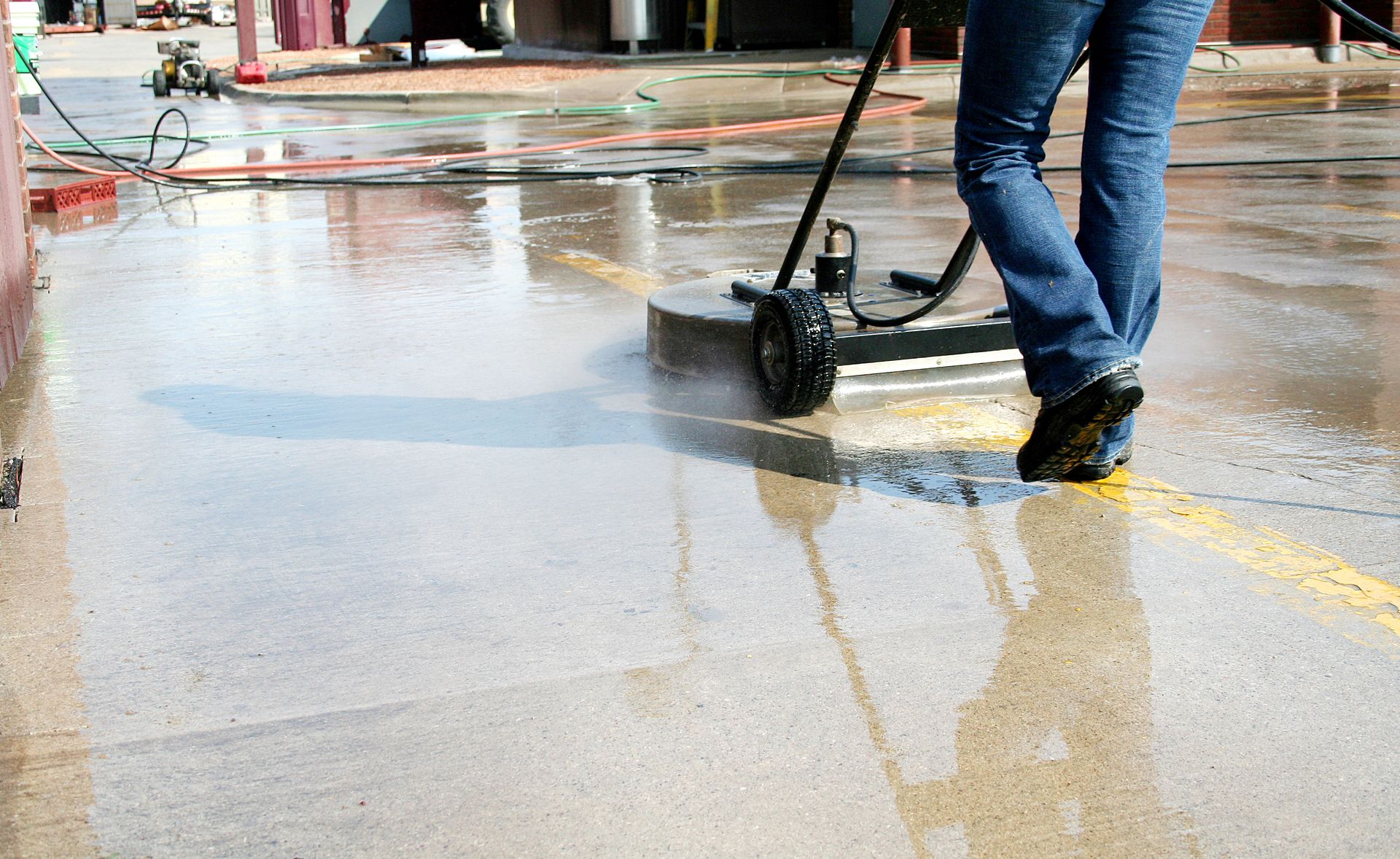 Person pressure-washing a wet sidewalk with a black machine, wearing jeans and yellow shoes.
