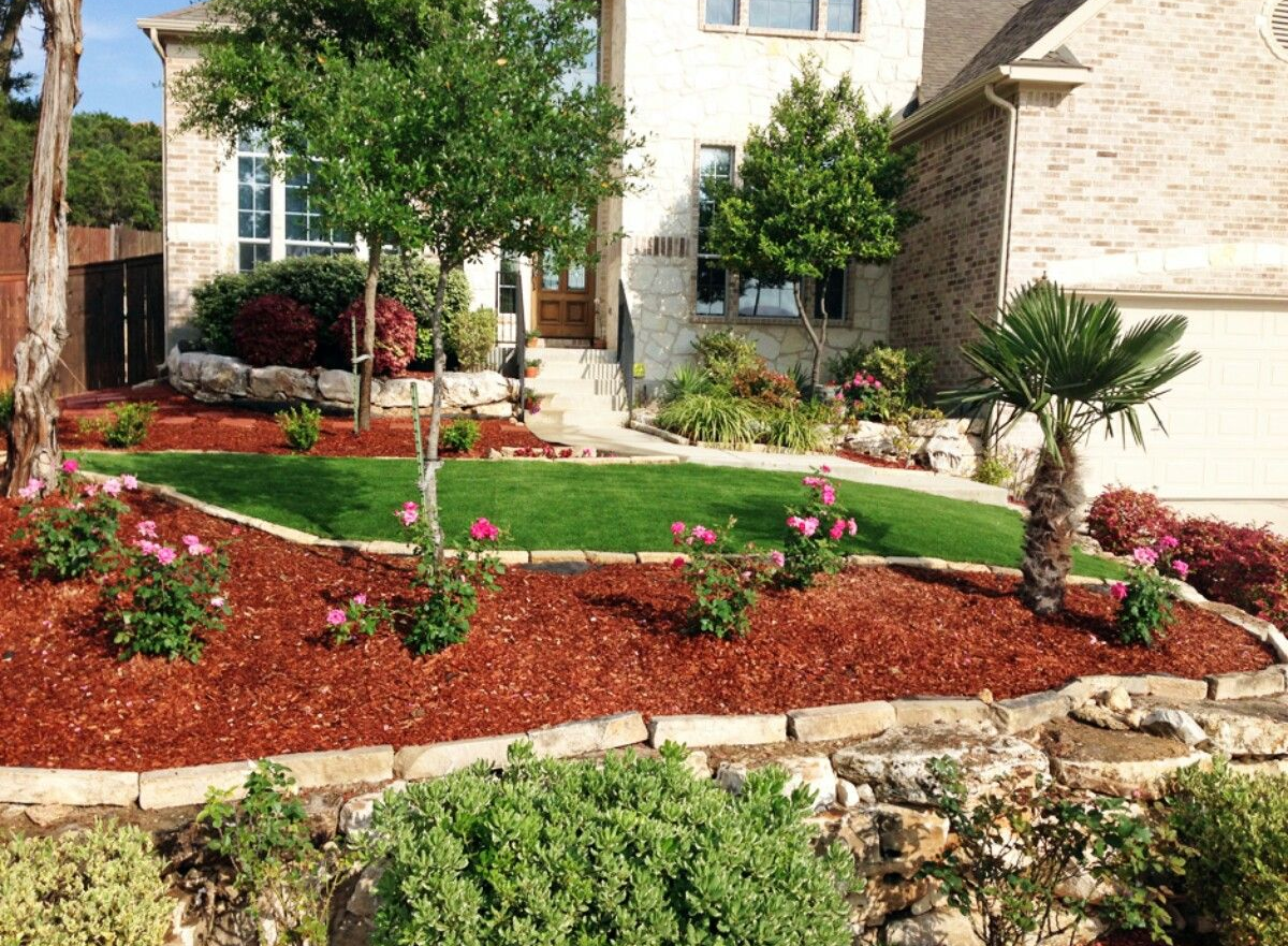 Landscaped front yard with green lawn, red mulch, pink flowers, and small trees beside a house