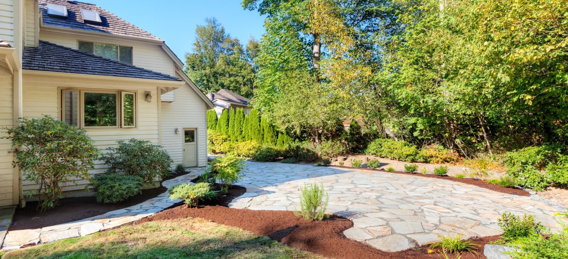 Exterior of a house with a stone patio, garden beds, and trees in bright sunlight