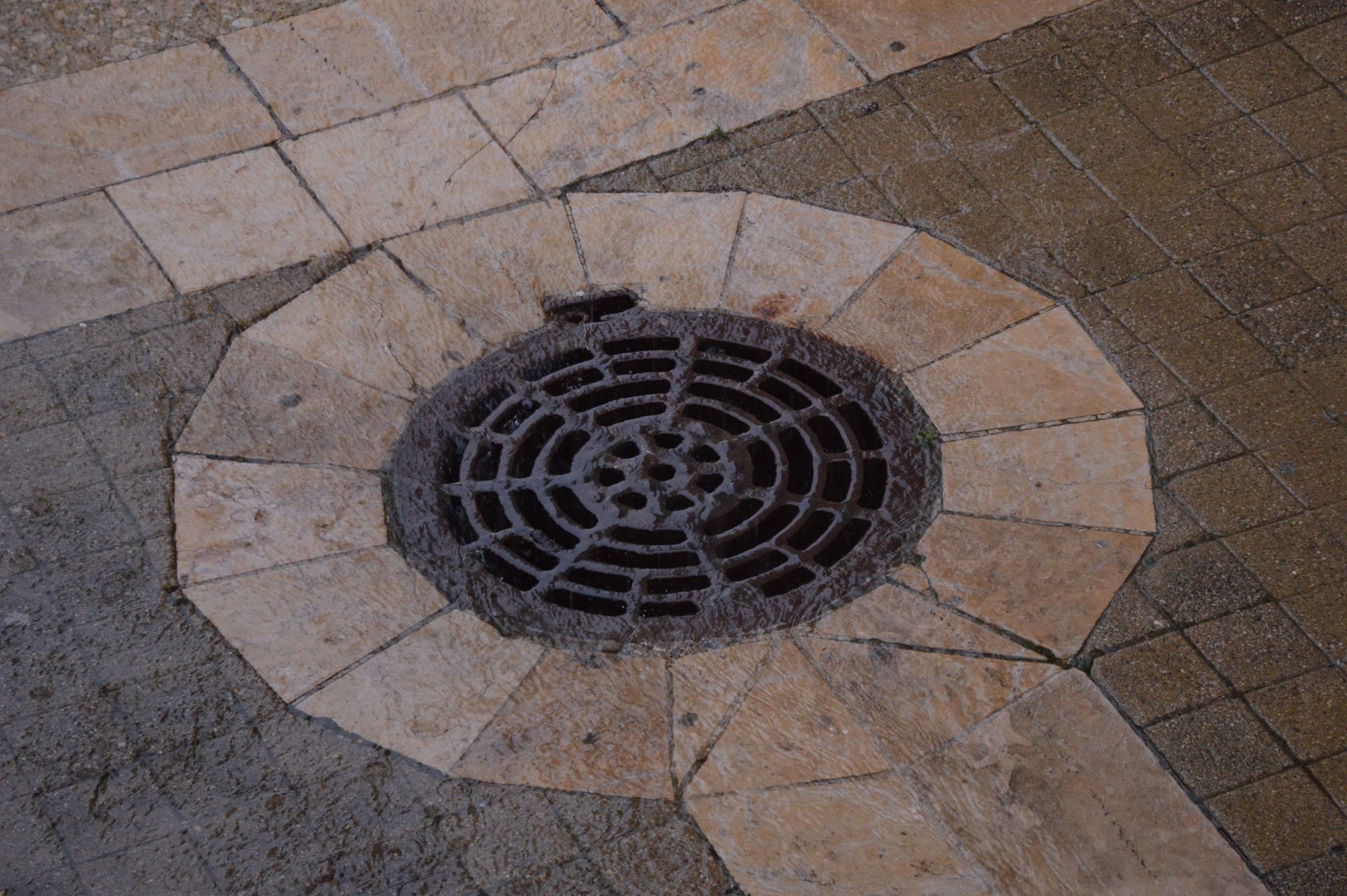 Round metal drain grate set in tan and brown stone pavement, viewed from above