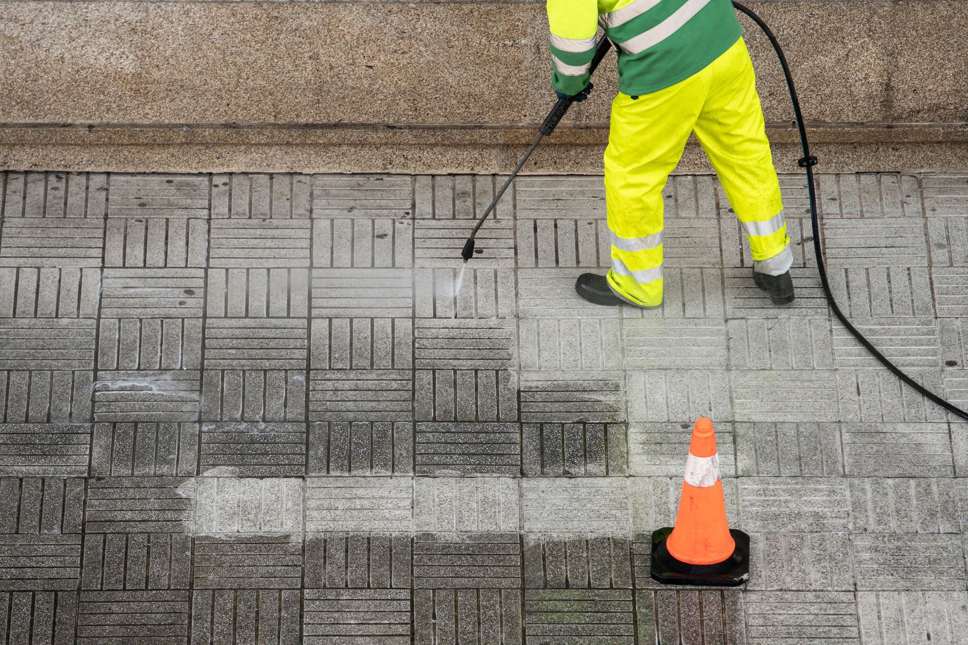 Worker pressure-washing patterned pavement beside an orange cone and hose