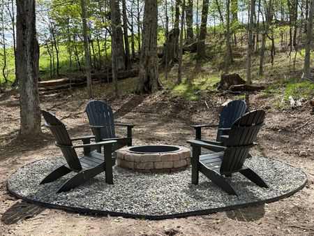 Stone fire pit with four black chairs on a gravel patio in a wooded backyard