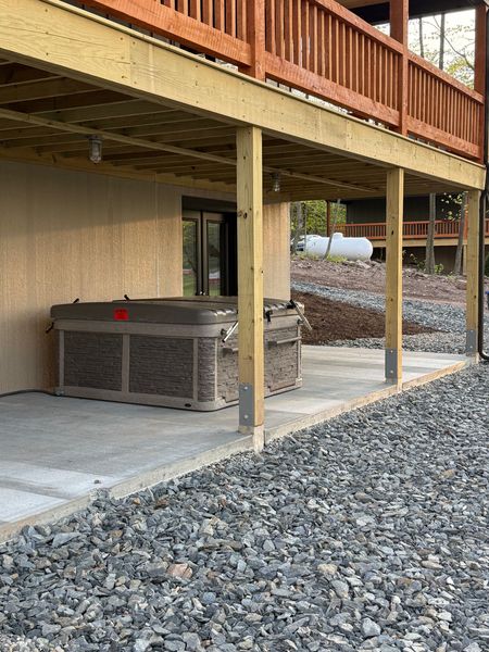 Covered patio under a wooden deck with support posts, concrete floor, and gravel landscaping