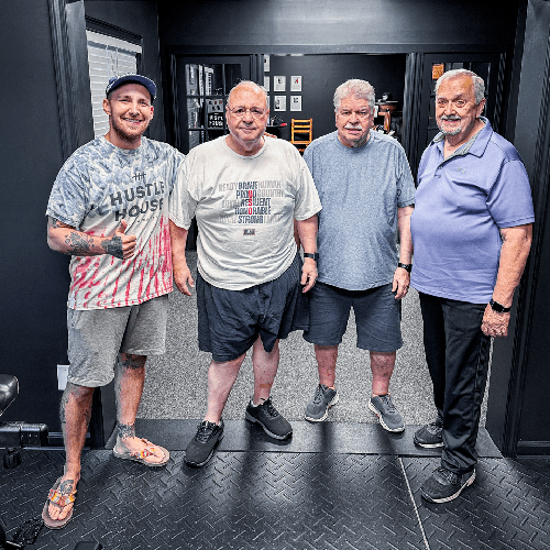 Four men standing in a gym, posing together in casual workout clothes.