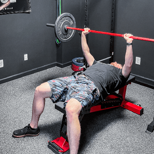 Man bench pressing a red barbell on a bench in a gym in Woodstock, GA, with weight plates and black flooring.