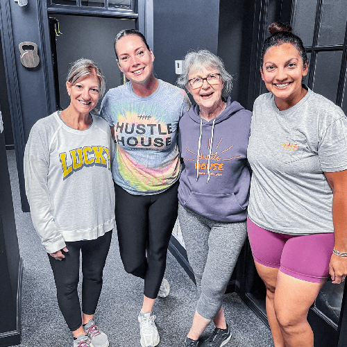 Four women in athletic clothes smiling together indoors, standing arm in arm.