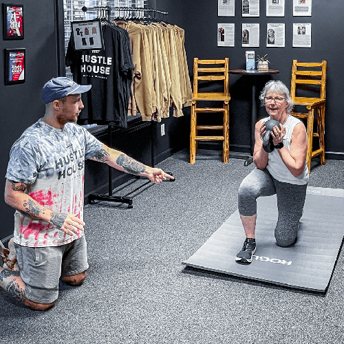 Two people exercising in a gym, one kneeling with weights and the other pointing while kneeling on a mat.