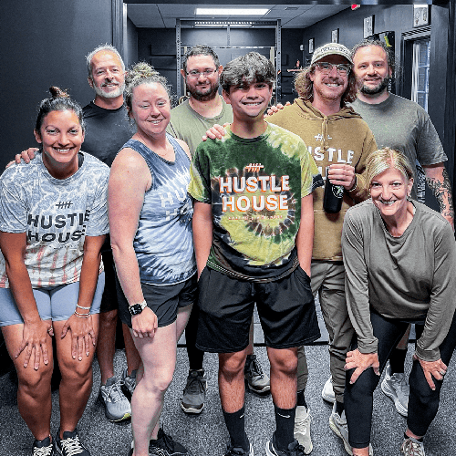 Group of smiling people posing indoors, several wearing “Hustle House” shirts.