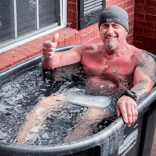 Man in a gray beanie relaxing in an icy stock tank, smiling and giving a thumbs-up beside a brick wall.