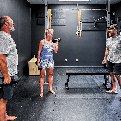 Three people standing in a gym, one lifting a dumbbell near a bench.