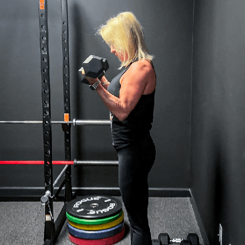 Person lifting a dumbbell in a gym, standing beside a rack and stacked weight plates.