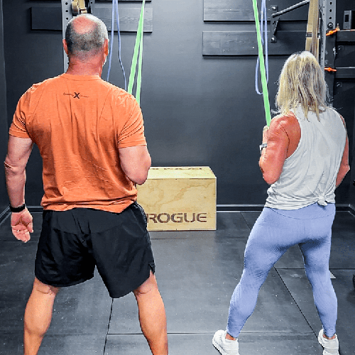 Two people exercising in a gym, standing with feet apart near a Rogue box.
