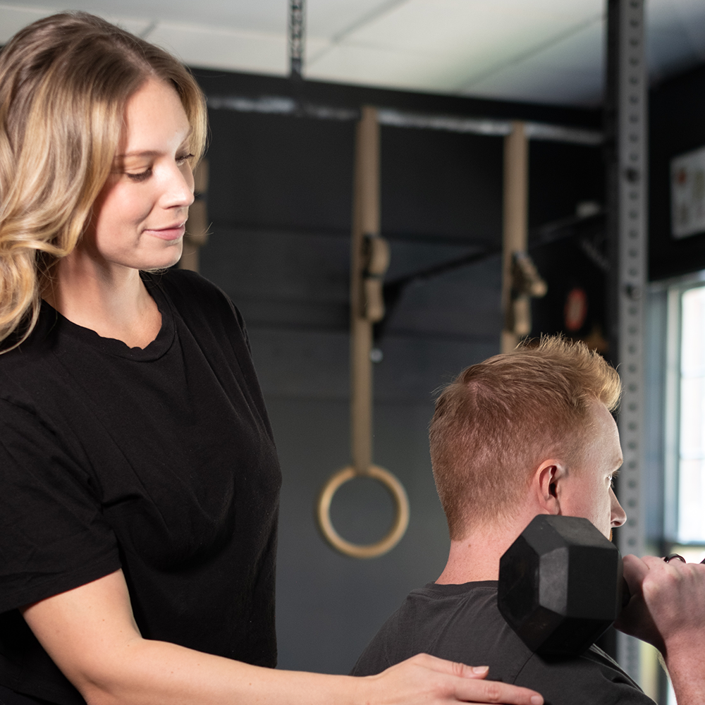Trainer coaching a person lifting a dumbbell in a gym, with gymnastic rings in the background