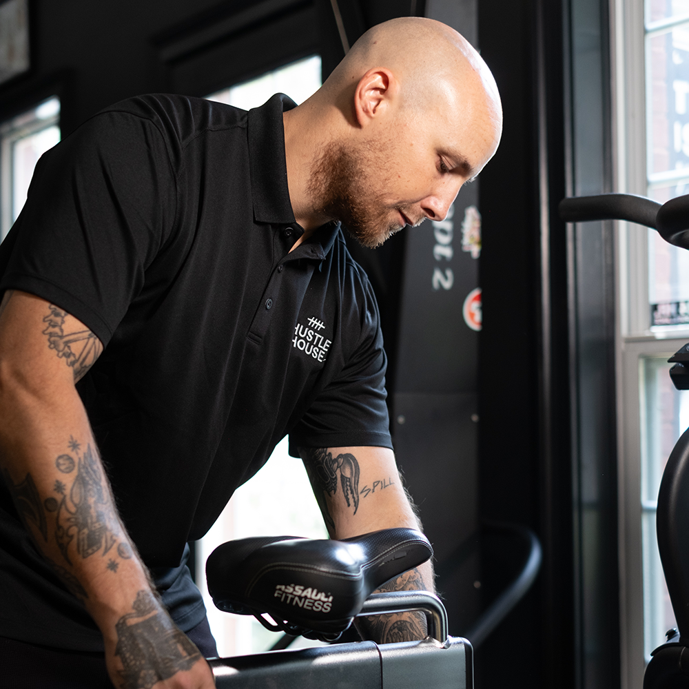 Person in a black polo leans over a gym machine near a window, focusing on the equipment.