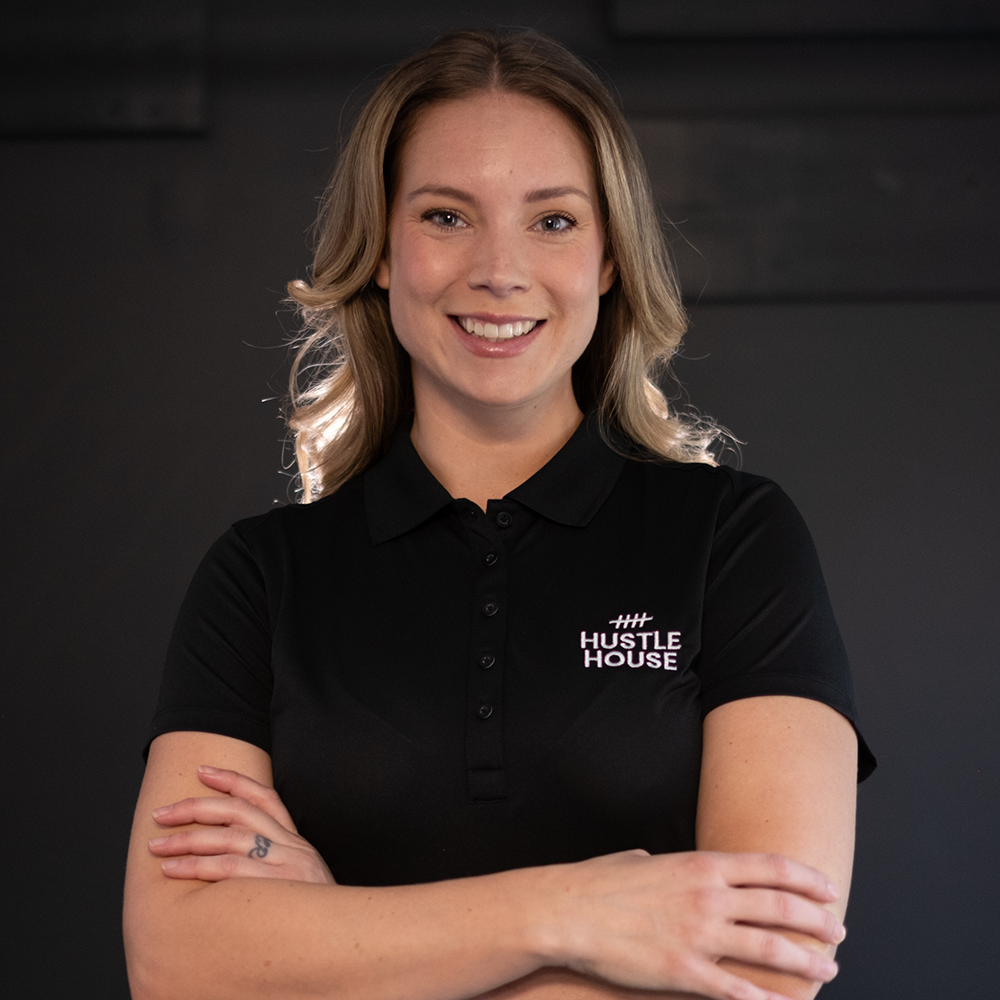 Smiling person in a black HUSTLE HOUSE polo, standing with arms crossed against a dark background