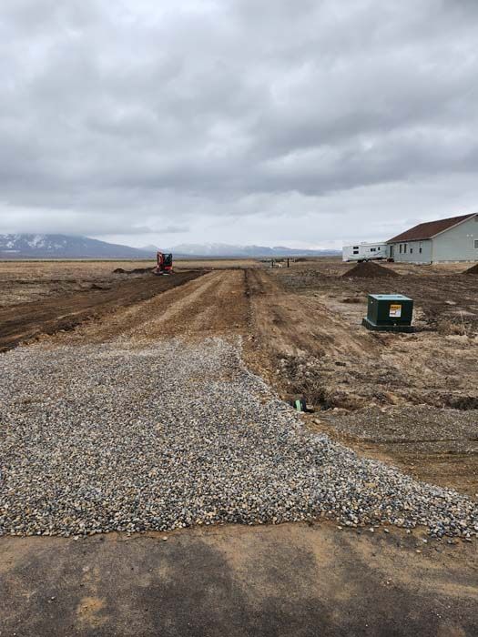 Gravel and dirt road under construction leading to buildings and a field under a cloudy sky.