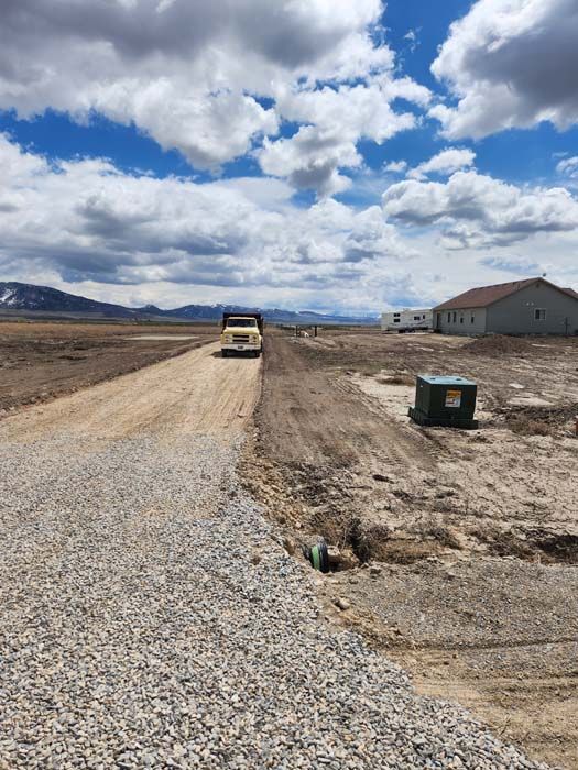 A gravel road with a truck, house, and utility box under a cloudy blue sky.
