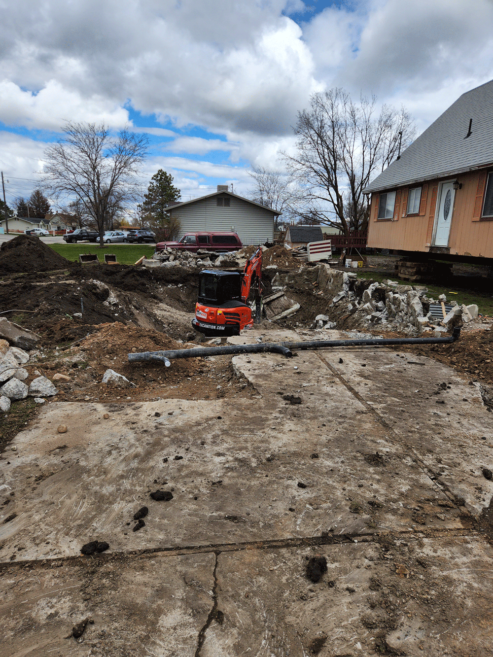 Construction site with orange excavator digging a muddy trench beside a house under cloudy skies