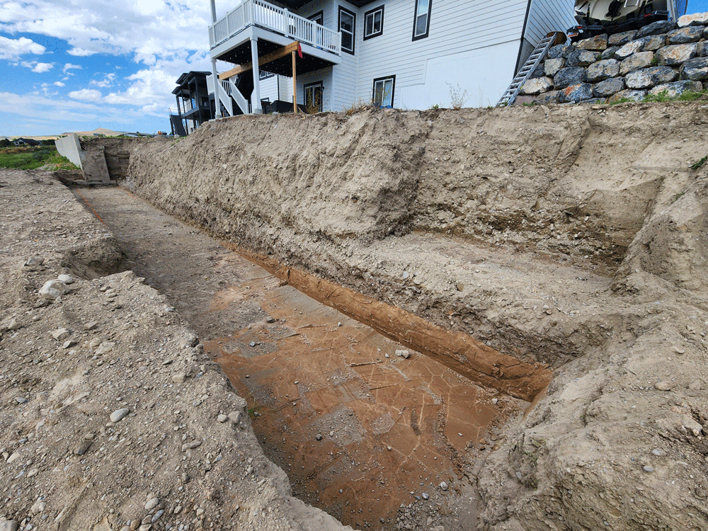 Excavated trench beside a white house on a rocky hillside under a partly cloudy sky