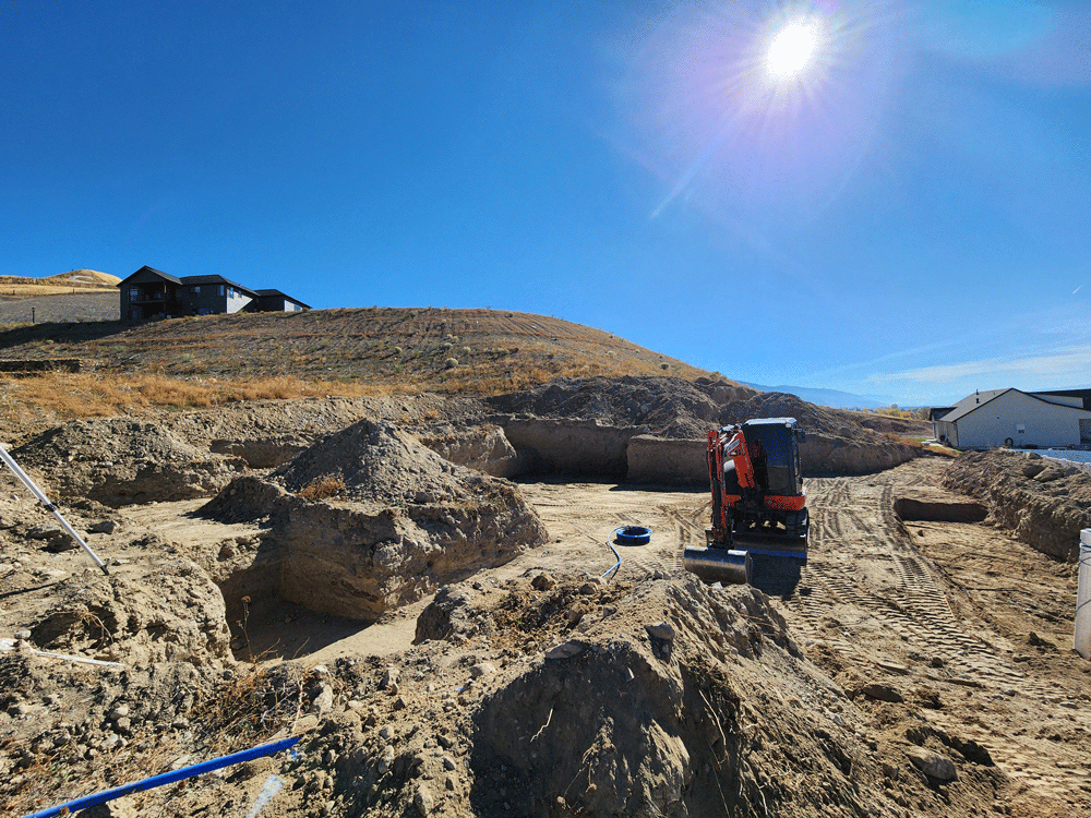 Excavator working on a sandy hillside under a bright blue sky, with houses in the background.