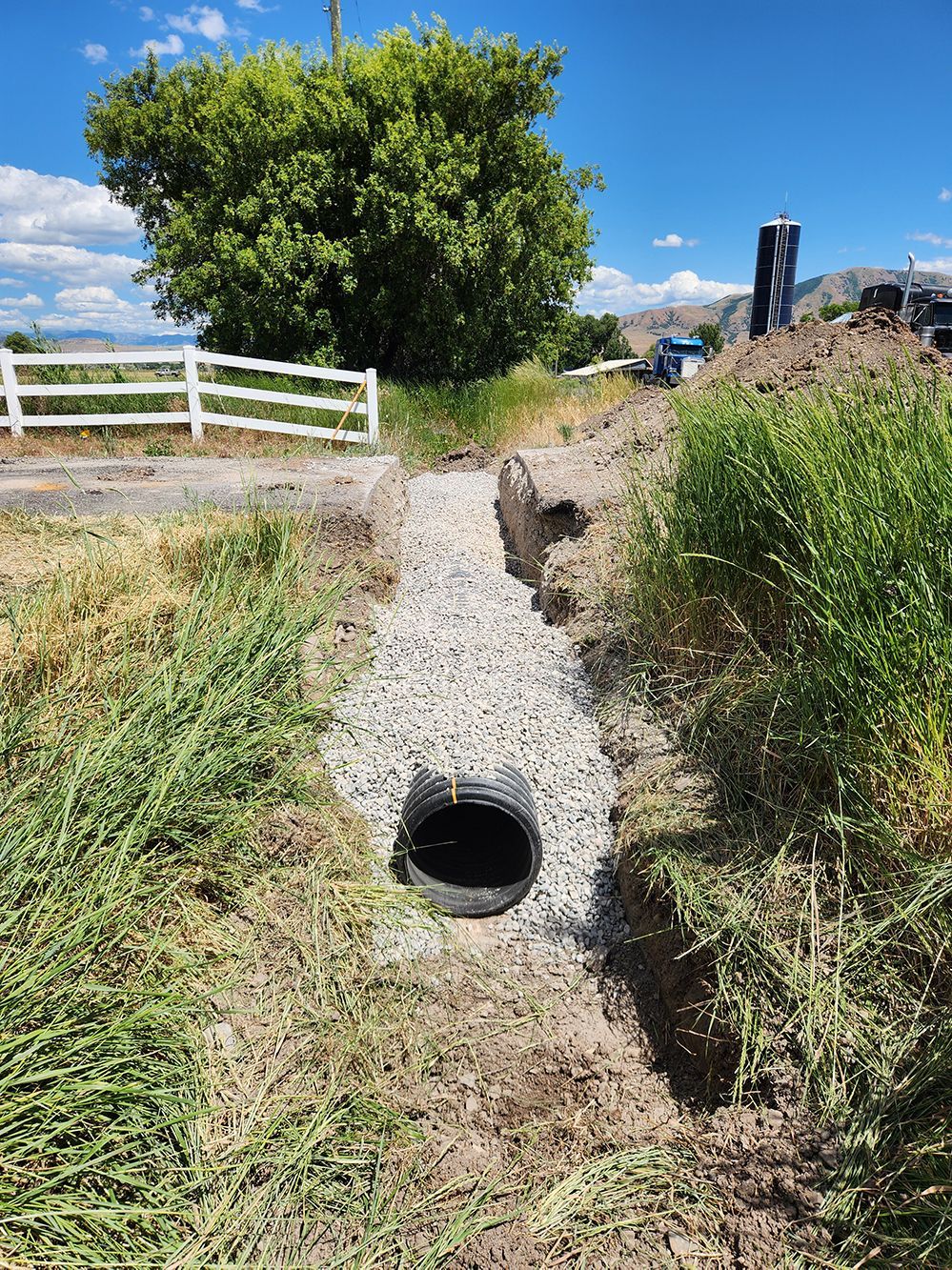 Gravel trench with a round drain opening beside a white fence and tall grass under a blue sky