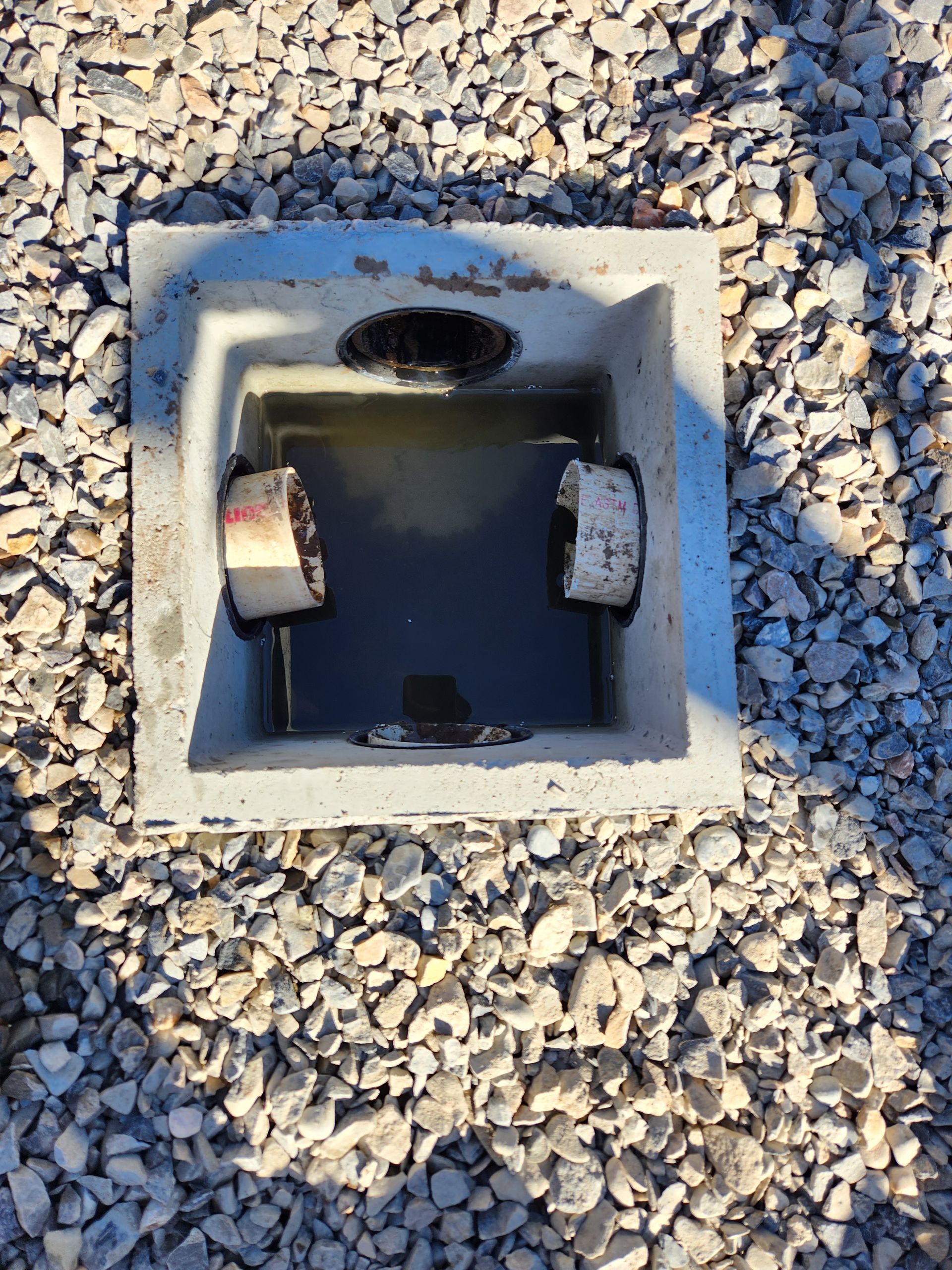 Square concrete utility box set in gravel, with a dark opening and two white components inside.